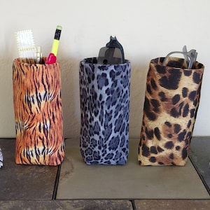 May include: Five fabric pen holders with animal print designs. The patterns include zebra, tiger, leopard, and a blue leopard print. Each holder contains pens, pencils, and glasses.