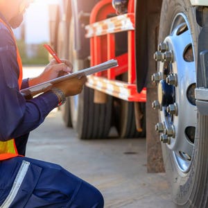 May include: A person in an orange safety vest and blue uniform kneels, writing on a clipboard next to a truck tyre. The tyre is black, and the wheel is silver with multiple lug nuts. The truck has a red and white safety bar.