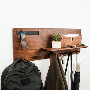 May include: A wooden wall-mounted organizer with a shelf, hooks, and a mail slot. It features a small potted plant, glasses, keys, and a jacket hanging on the hooks.