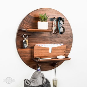 May include: A round wooden wall organizer with shelves, hooks, and a mail slot. It features a small potted plant, sunglasses, keys, and a jacket hanging on the hooks.