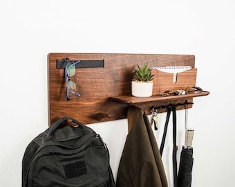May include: A wooden wall-mounted organizer with a shelf, hooks, and a mail slot. It features a small potted plant, glasses, keys, and a jacket hanging on the hooks.