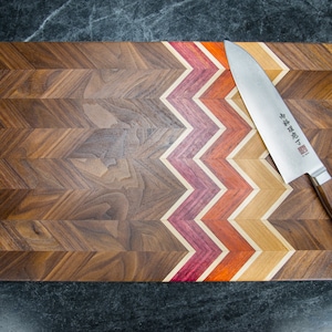Może przedstawiać: Drewniana deska do krojenia z wzorem w chevron. Deska wykonana jest z drewna orzechowego z kontrastową inkrustacją z czerwonego, pomarańczowego i ż&oacute;łtego drewna. Srebrny n&oacute;ż z drewnianym uchwytem leży na desce. N&oacute;ż ma japoński tekst na ostrzu.