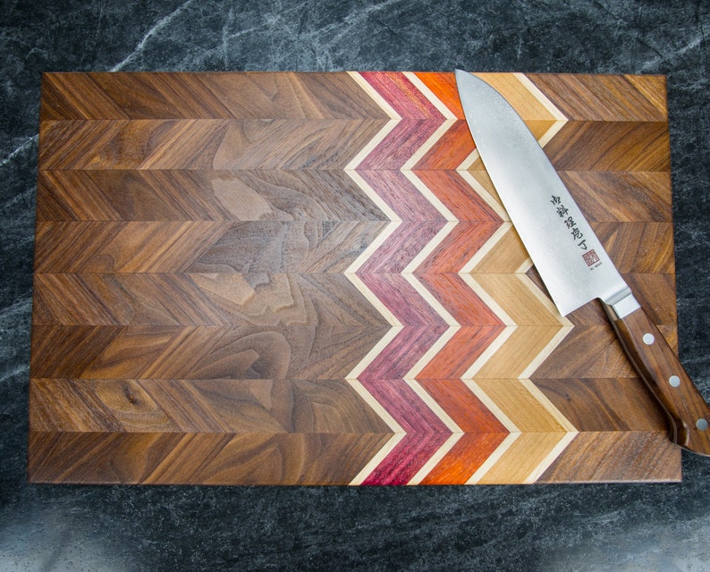 Może przedstawiać: Drewniana deska do krojenia z wzorem w chevron. Deska wykonana jest z drewna orzechowego z kontrastową inkrustacją z czerwonego, pomarańczowego i ż&oacute;łtego drewna. Srebrny n&oacute;ż z drewnianym uchwytem leży na desce. N&oacute;ż ma japoński tekst na ostrzu.