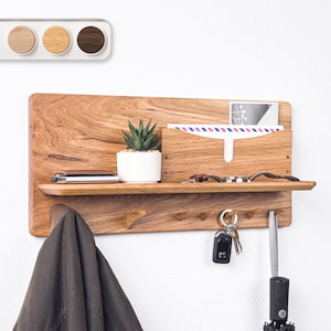 May include: A wooden wall-mounted organizer with a shelf, hooks, and a mail slot. The organizer is made of light brown wood with a natural finish. It has a small potted succulent plant on the shelf, a jacket hanging on a hook, and keys and an umbrella hanging on other hooks.