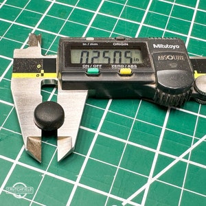 May include: A black digital caliper measuring a small black round object. The caliper reads 0.2505 inches.