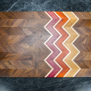 Może przedstawiać: Drewniana deska do krojenia z wzorem w chevron. Deska wykonana jest z drewna orzechowego z kontrastową inkrustacją z drewna czerwonego, pomarańczowego i brązowego.
