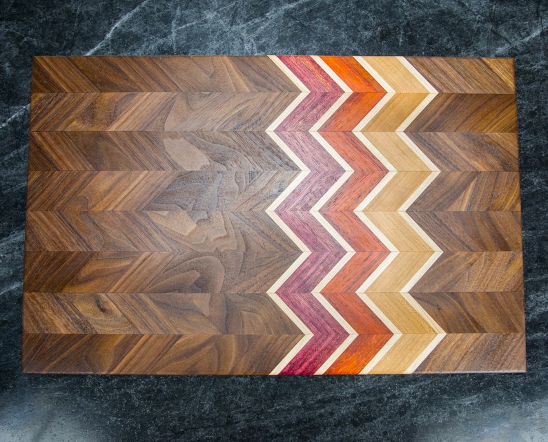 Może przedstawiać: Drewniana deska do krojenia z wzorem w chevron. Deska wykonana jest z drewna orzechowego z kontrastową inkrustacją z drewna czerwonego, pomarańczowego i brązowego.