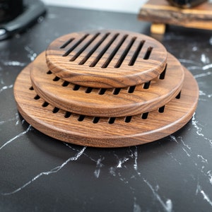 May include: Three round wooden coasters with a slatted design, stacked on top of each other. The coasters are made of dark brown wood and have a natural finish.