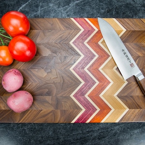 Może przedstawiać: Drewniana deska do krojenia z chevronowym wzorem w odcieniach brązu, czerwieni i ż&oacute;łci. Na desce leży n&oacute;ż, a na niej znajdują się pomidory i ziemniaki.