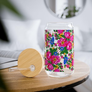 May include: A glass can with a floral pattern in pink, blue, and green. The can has a bamboo lid with a hole for a straw. The straw is clear.