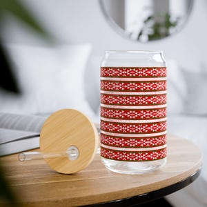 May include: A clear glass drinking cup with a bamboo lid and straw. The cup features a repeating pattern of red, white, and green geometric shapes. The cup is sitting on a wooden table.