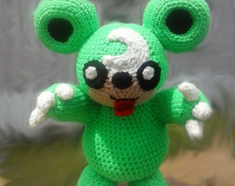 May include: A handmade green crochet creature with large round ears, black eyes, and a white question mark on its head. The plush toy has white paws and a red tongue, giving it a playful appearance. The background is a soft, blurred gradient.