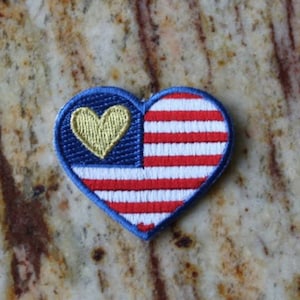 May include: A red, white, and blue heart-shaped patch with a gold heart in the center. The patch is embroidered with the American flag.