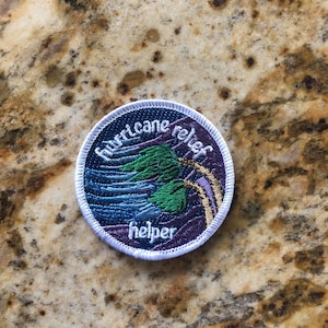 May include: A white, round embroidered patch with a purple background and the text "hurricane relief helper" in white. The patch features a stylized image of a palm tree and waves in blue, green, and gold.