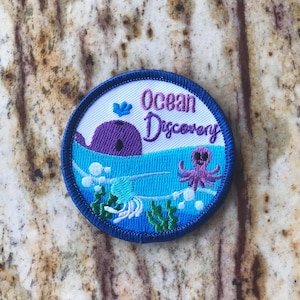 May include: A blue and white embroidered patch with the text "Ocean Discovery". The patch features a purple whale, a pink octopus, and blue and green seaweed.
