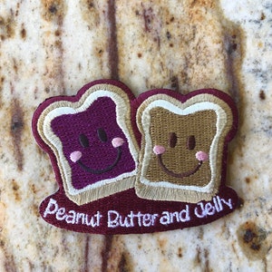 May include: Embroidered patch featuring two smiling slices of bread, one with purple jelly and the other with peanut butter. The patch has a maroon border and the words "Peanut Butter and Jelly" in white.