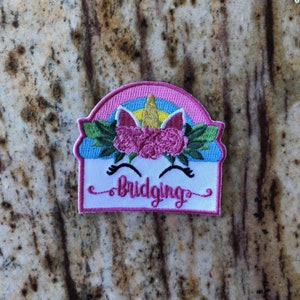 May include: A white embroidered patch with a pink, blue, and gold unicorn design. The unicorn has a rainbow mane and a flower crown. The text "Bridging" is embroidered in pink on the patch.