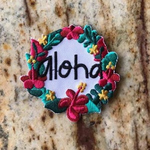 May include: A white embroidered patch with a floral wreath surrounding the word "Aloha". The flowers are red, pink, yellow, and green.