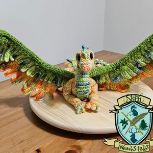 May include: A crocheted dragon with green, orange, and yellow wings. The dragon is sitting on a wooden base with a logo that says "Steph Weaves Tales".