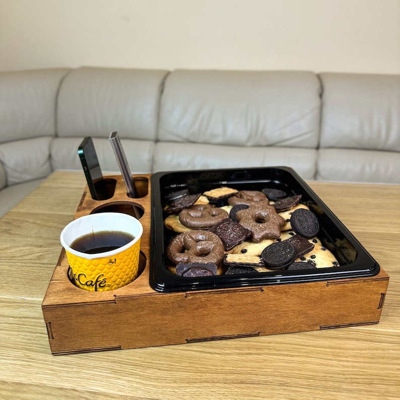 May include: A wooden serving tray with a black tray insert for holding snacks. The tray has a built-in cup holder and a separate compartment for a phone or other small items. The tray is filled with a variety of chocolate chip and shortbread cookies.
