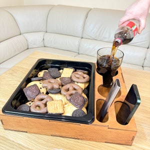 May include: A wooden tray with compartments for snacks and drinks. The tray has a black insert with a variety of cookies and a glass of cola. There is also a phone and a silver utensil in the tray.