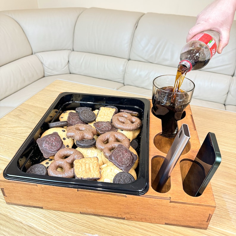 May include: A wooden tray with compartments for snacks and drinks. The tray has a black insert with a variety of cookies and a glass of cola. There is also a phone and a silver utensil in the tray.