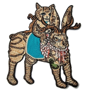 Może przedstawiać: Naszywka haftowana przedstawiająca brązowo-białego królika z rogami, noszącego czerwony i biały naszyjnik z koralików. Na grzbiecie królika siedzi brązowo-biały kot.