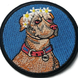 Puede incluir: Un parche bordado circular que muestra un perro marrón con una corona de margaritas y un collar rojo con detalles azules. El perro tiene una nariz rosa y una pequeña placa circular plateada colgando del collar sobre un fondo azul.