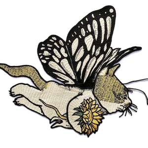 Op de afbeelding: Geborduurde patch met een kat met vlindervleugels en een zonnebloem. De kat is crème en goud, met zwart-witte vlindervleugels. De zonnebloem is geel en groen. De patch is bedoeld om op te strijken.