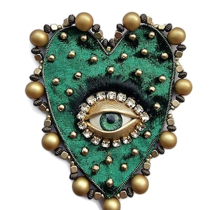 May include: A heart-shaped brooch with a green velvet base, decorated with gold beads and a central eye design. The eye has a green iris, surrounded by clear crystals and a gold frame. A unique accessory.