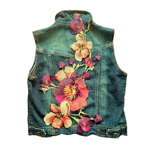 May include: A denim vest with a floral design. The vest is a medium wash blue with a colourful embroidered floral pattern on the back. The flowers are shades of pink, purple, and yellow.
