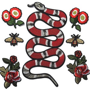 May include: A collection of embroidered patches featuring a large central snake with red, white, and black bands, two bees with gold wings and striped bodies, and four floral arrangements with red petals and green leaves on a plain background.