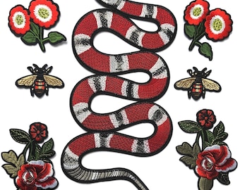 Op de afbeelding: Een verzameling geborduurde patches met een grote centrale slang met rode, witte en zwarte banden, twee bijen met gouden vleugels en gestreepte lichamen, en vier bloemstukken met rode bloemblaadjes en groene bladeren op een effen achtergrond.