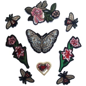 Puede incluir: Un conjunto de parches bordados para planchar que incluyen una mariposa, flores y abejas. Los parches son en tonos de rojo, rosa, verde, blanco y negro.