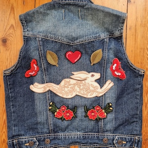 Puede incluir: Un chaleco vaquero azul adornado con parches bordados. Los parches incluyen un conejo corriendo, mariposas rojas, un corazón rojo, hojas verdes y flores rojas. El chaleco tiene una parte delantera abotonada y un cuello clásico.
