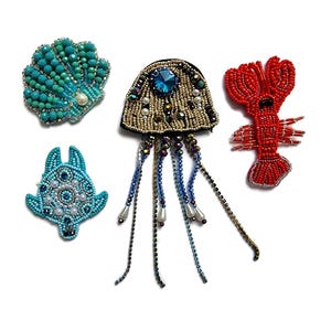 May include: A selection of beaded ocean-themed brooches. The brooches feature a turquoise seashell, a blue turtle, a jellyfish with blue and gold beads, and a red lobster. Each brooch is intricately detailed with various bead sizes and colours.