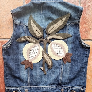 May include: A denim vest with a decorative embroidered design of two light-colored fruits with a brown stem and leaves. The fruits have a white grid pattern. The vest is a medium blue color.