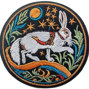 Op de afbeelding: Geborduurde strijkpatch met een wit konijn met een rode en gouden halsband, omringd door groen gebladerte, een halve maan en sterren op een zwarte achtergrond.