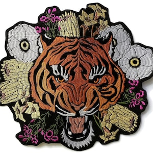 May include: Embroidered patch of a tiger's head with orange and black stripes, framed by white, yellow, and pink floral details. The patch has a black border and is suitable for clothing or accessories.