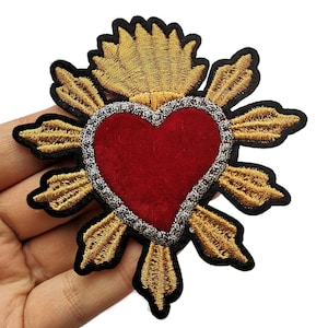 Op de afbeelding: Een rood fluwelen hart met gouden geborduurde vleugels en een gouden vlam bovenop. Het hart is omgeven door een zilveren parelrand.