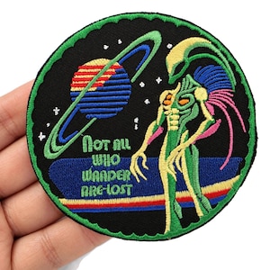 May include: A circular embroidered patch with a black background featuring a green alien figure, a striped planet with rings, and white stars. The text "Not all who wander are lost" is stitched in green lettering above a blue and red horizon line.