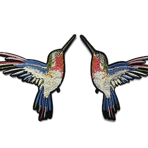 Puede incluir: Dos parches bordados de colibrí con alas coloridas y un contorno negro. Los colibríes se miran con los picos apuntando hacia arriba.