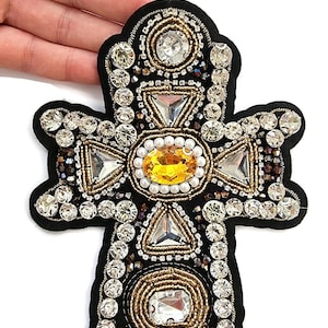 May include: Black cross-shaped embellishment with a detailed design. The cross features a central oval yellow gem, surrounded by clear and gold-toned crystals, and triangular gems. The edges are outlined with gold beading.