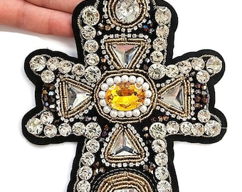 May include: Black cross-shaped embellishment with a detailed design. The cross features a central oval yellow gem, surrounded by clear and gold-toned crystals, and triangular gems. The edges are outlined with gold beading.