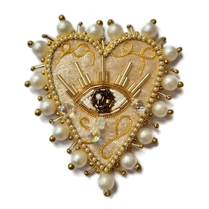 Peut inclure: Broche en forme de cœur de couleur crème avec un motif d'œil brodé. La broche est ornée de perles dorées, de perles et de cristaux. L'œil est détaillé avec des perles blanches, marron et dorées, et le cœur est souligné de fil d'or.