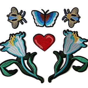 May include: Embroidered patches featuring a butterfly, two bees, two flowers, and a red heart. The butterfly has blue and brown wings. The bees are gold and blue. The flowers are blue and white with yellow accents. The patches have black borders.