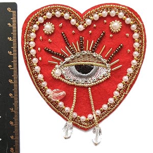 May include: A red heart-shaped appliqué with an embroidered eye design. It's adorned with gold beads, pearls, and clear crystal teardrop pendants. The eye has detailed embroidery with gold and silver accents.