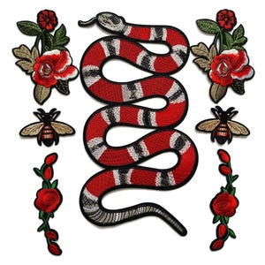 Pode incluir: Patches bordados com uma cobra vermelha e branca, rosas vermelhas e abelhas. A cobra é o design central, com um corpo vermelho e listras brancas. Os patches são provavelmente para roupas ou acessórios.