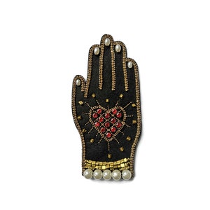 May include: Black hand-shaped patch with gold and red embellishments. The patch features a heart design made of red gems and is outlined with gold beads. The fingertips have pearl accents, and the base has a row of pearls.