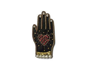 Puede incluir: Parche negro con forma de mano con adornos dorados y rojos. El parche presenta un diseño de corazón hecho de gemas rojas y está delineado con cuentas doradas. Las puntas de los dedos tienen detalles de perlas y la base tiene una fila de perlas.